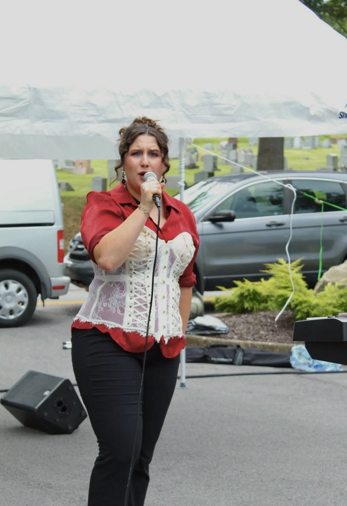 A woman in a red shirt and white corset-style top speaking into a microphone at an outdoor event. There are parked cars and a cemetery with headstones in the background.