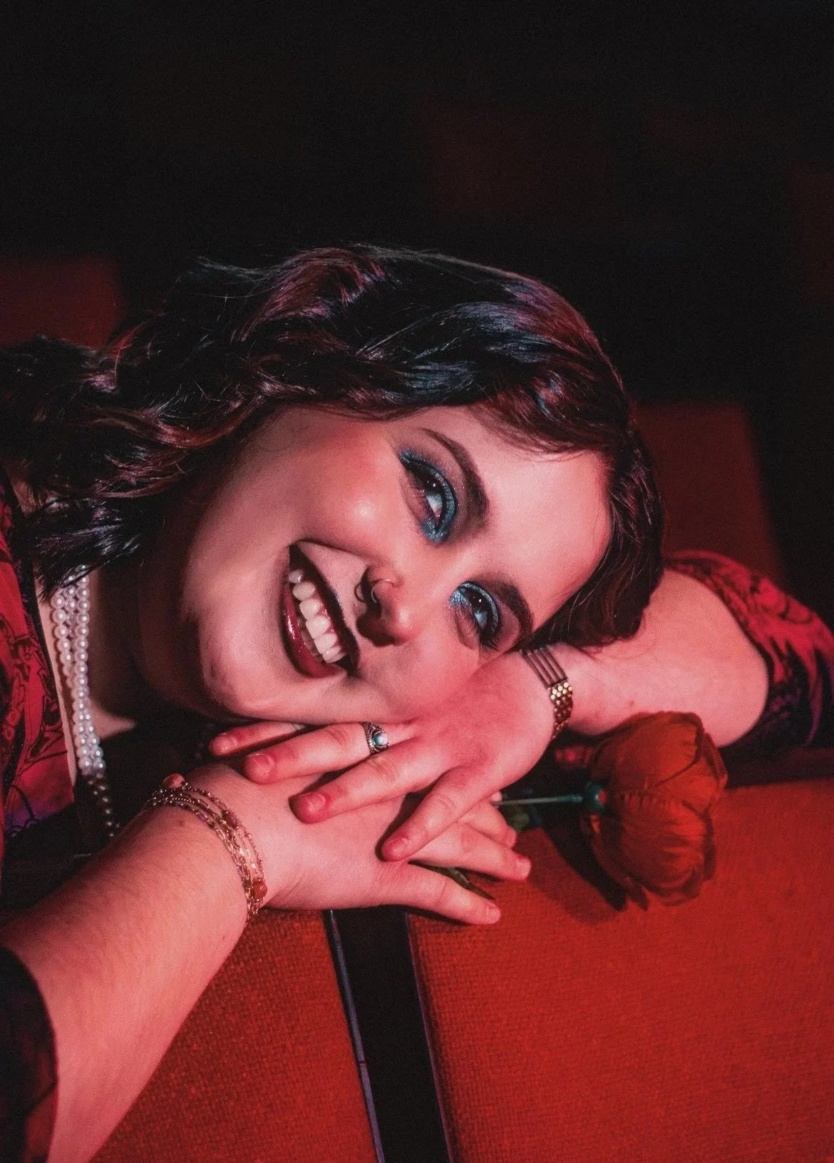 A woman with dark hair styled in waves, wearing blue eyeshadow, smiling with her hands resting on a red surface. She has jewelry including rings, a bracelet, a watch, and a pearl necklace. A single red rose is on the surface near her hands.