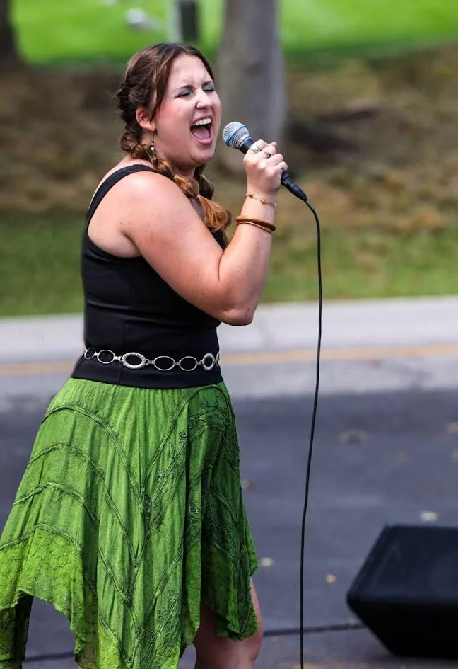 A woman singing passionately into a microphone outdoors, wearing a black sleeveless top and a green skirt.