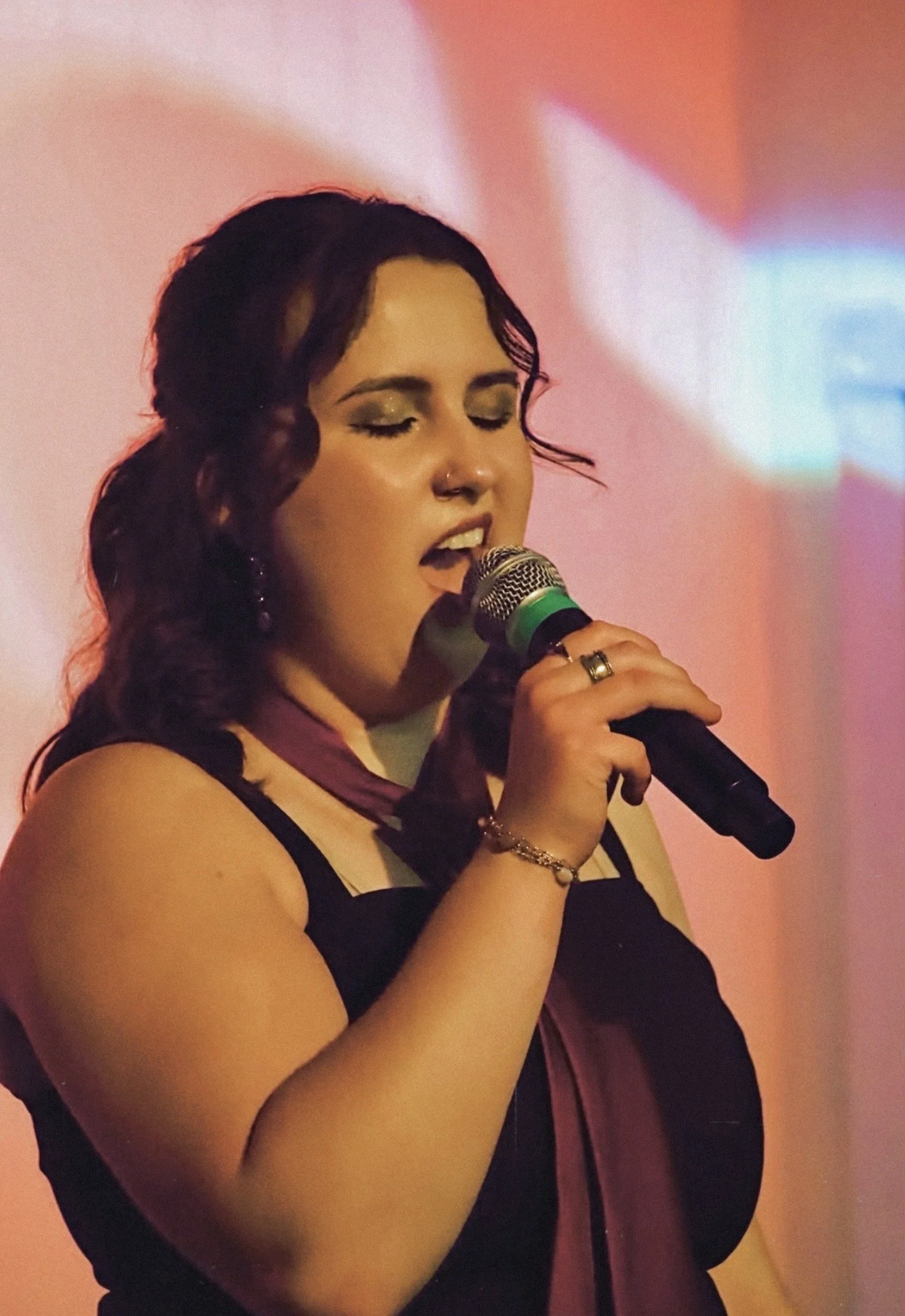 A young woman with dark wavy hair, makeup, and a nose piercing singing into a microphone against a pink and orange background.