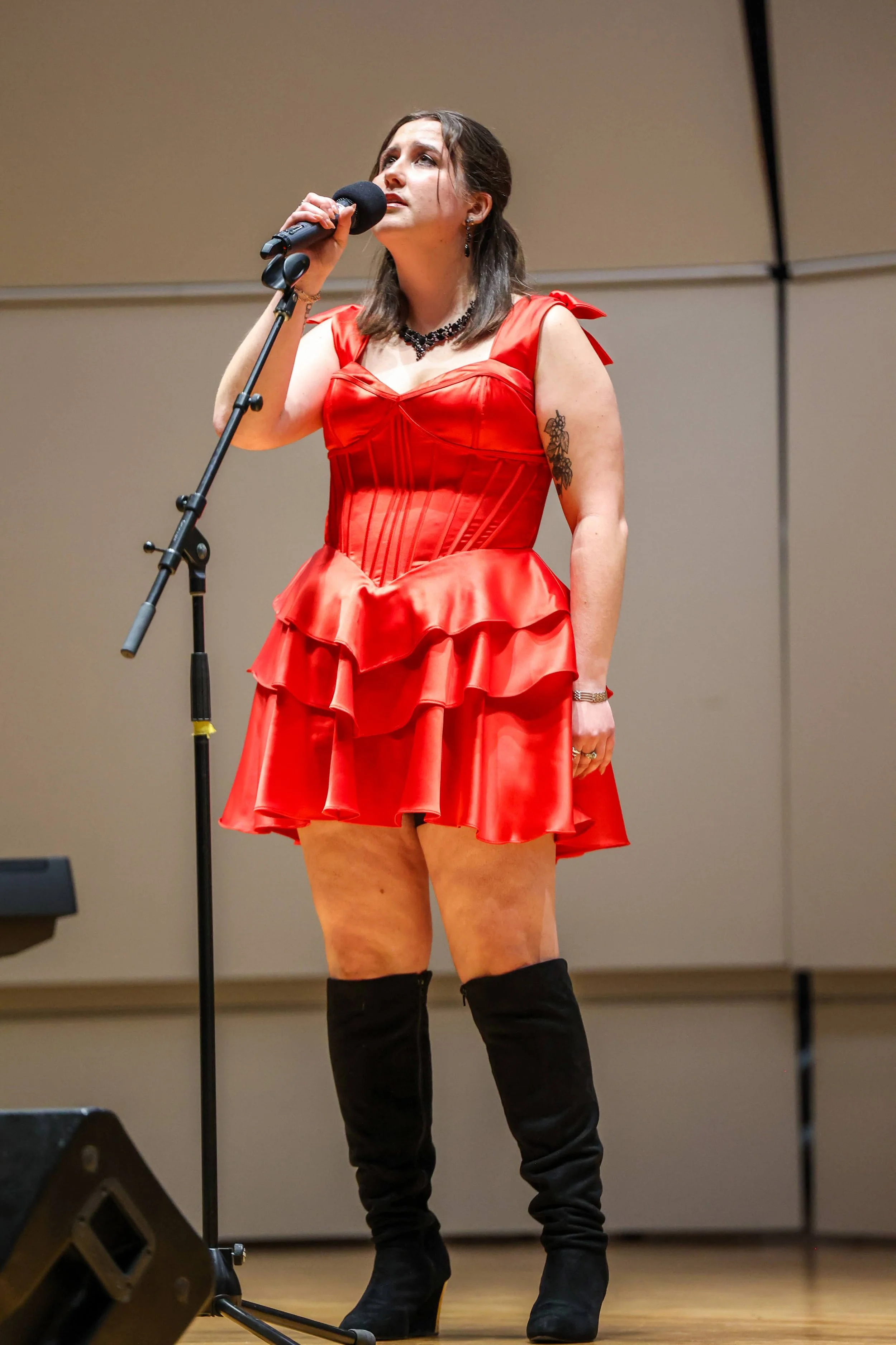 A woman wearing a red dress and black thigh-high boots stands on stage holding a microphone, looking upward.