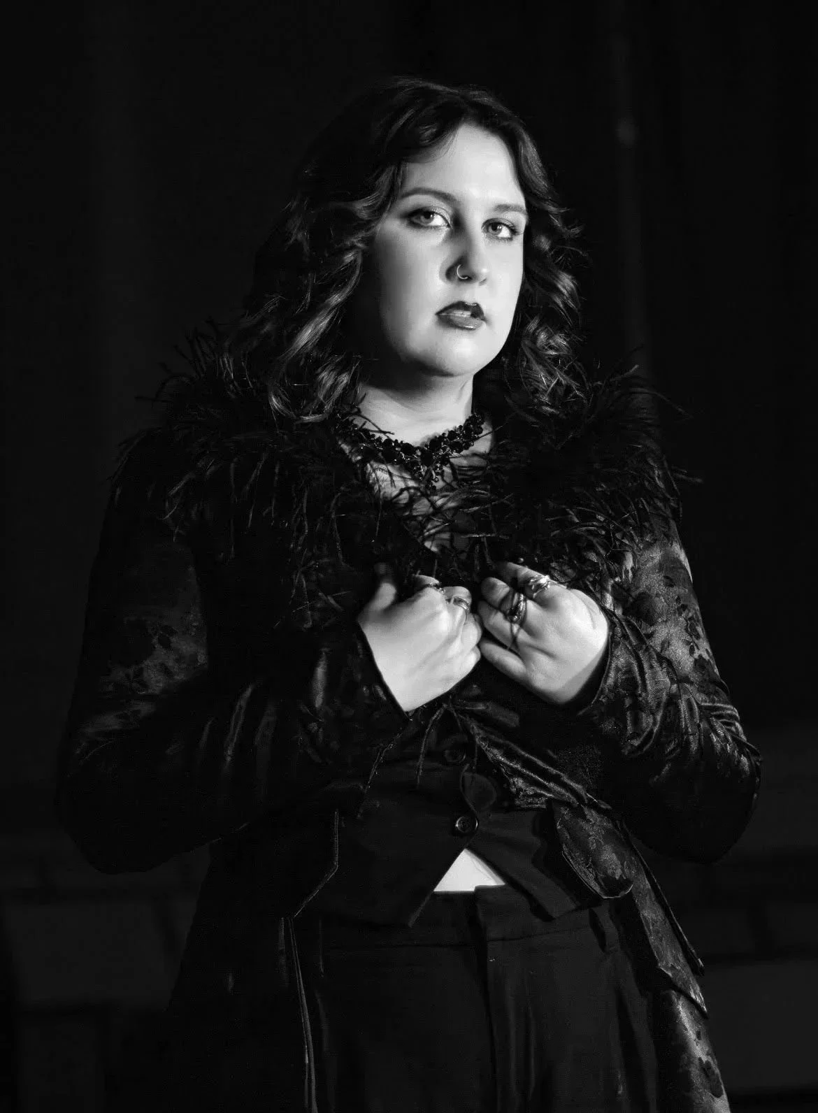 A woman with dark, curly hair and a nose ring wearing a black, textured dress with feathered shoulders and a necklace, posing with her hands on her chest against a dark background.