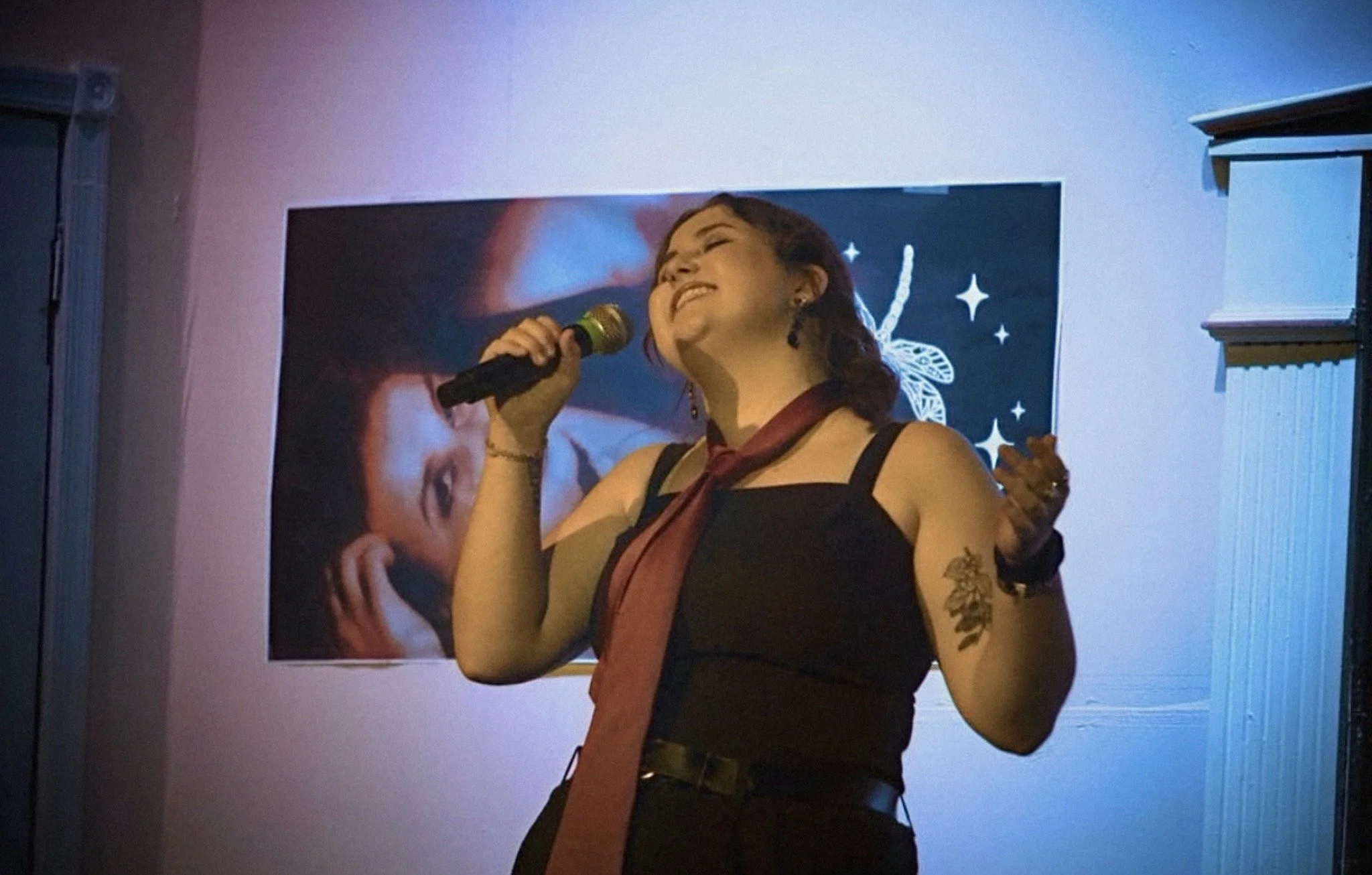 A woman with short brown hair singing into a microphone, with a background featuring a butterfly design and a large mural or poster of a woman.