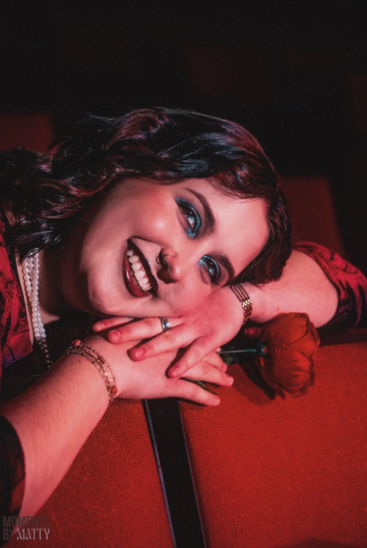 Woman with dark hair and blue makeup, smiling and resting her head on her hand, holding a red rose, wearing jewelry including rings, bracelets, and a pearl necklace, with a dark background.