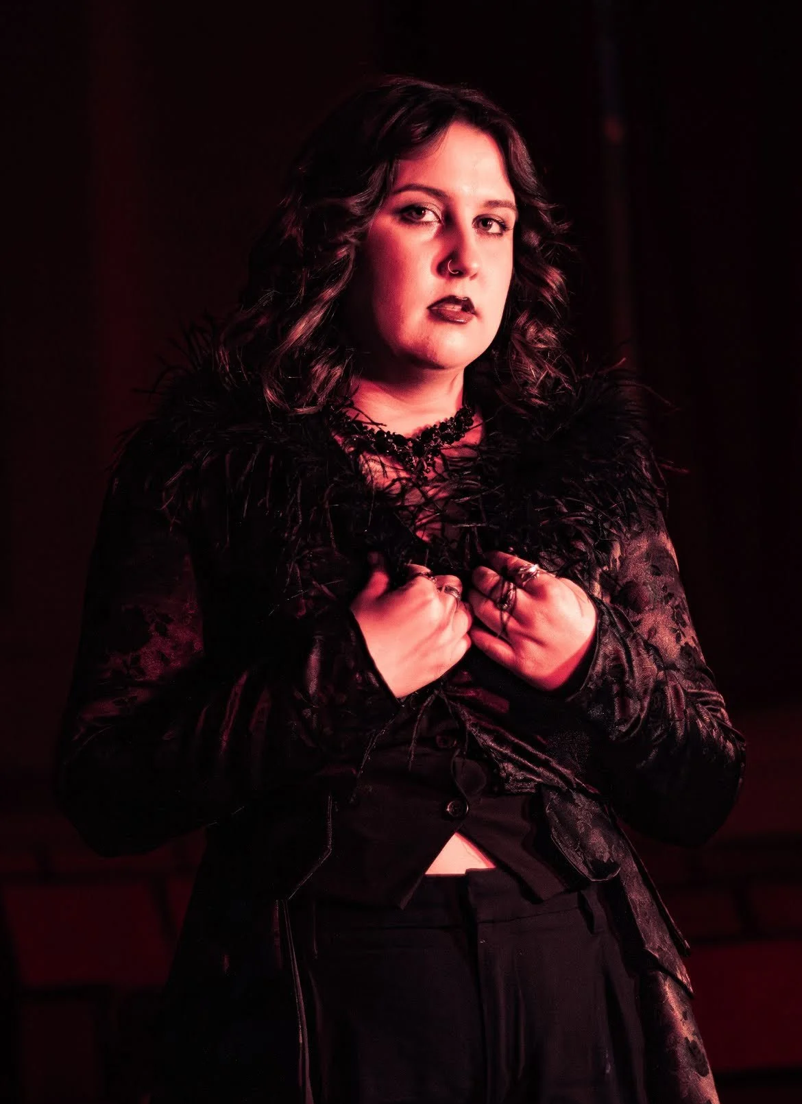 A person with dark hair and makeup, wearing a black feathered jacket and jewelry, posing against a dark red background.