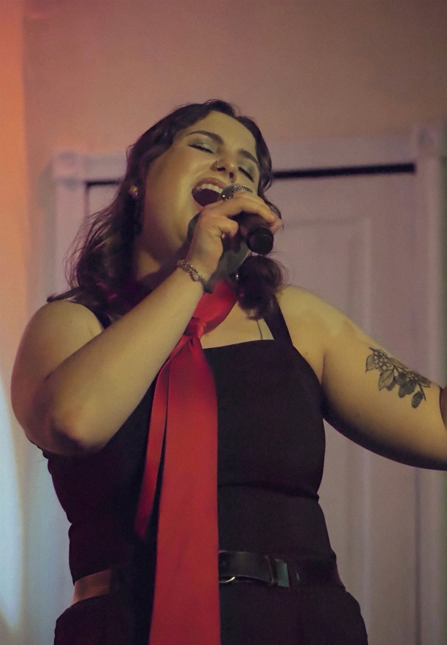 A woman singing into a microphone, with her eyes closed and mouth open, wearing a black dress with red accents, a necklace, and a tattoo on her left arm.