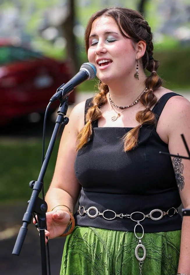 Young woman with braided hair singing into a microphone outdoors, wearing a black top and green skirt.