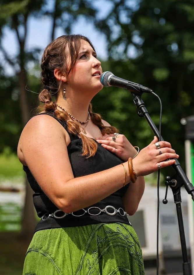 Young woman singing or speaking into a microphone outdoors, with trees in the background.