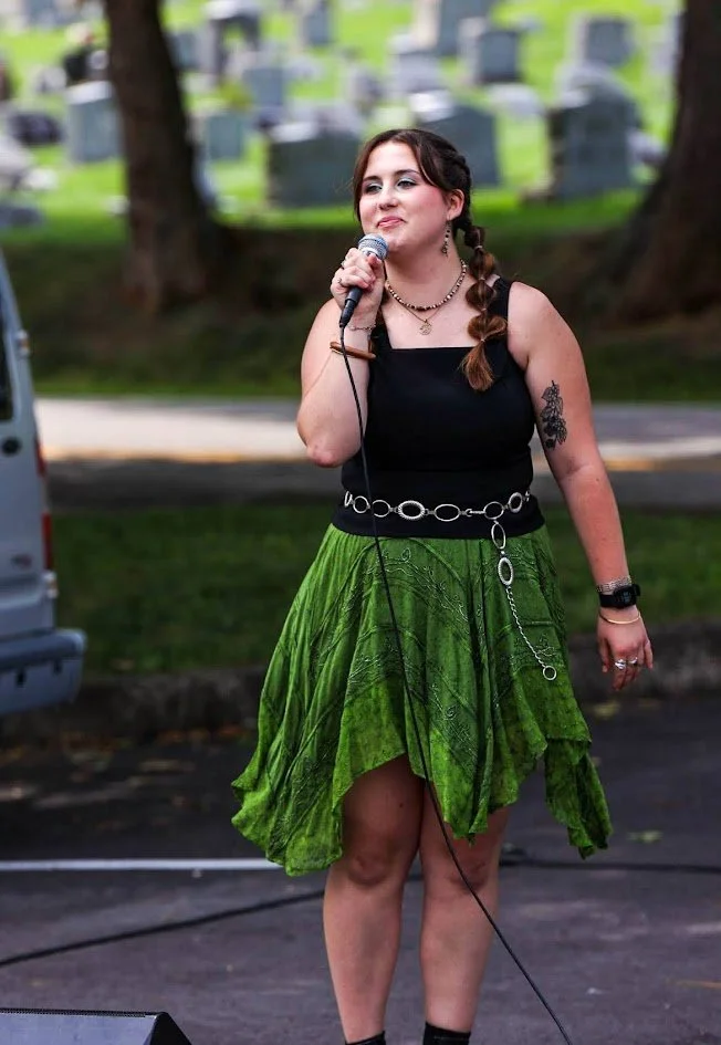 A young woman with braided hair stands outdoors holding a microphone. She is wearing a black sleeveless top, a green skirt, and a chain belt. Behind her is a cemetery with tombstones.