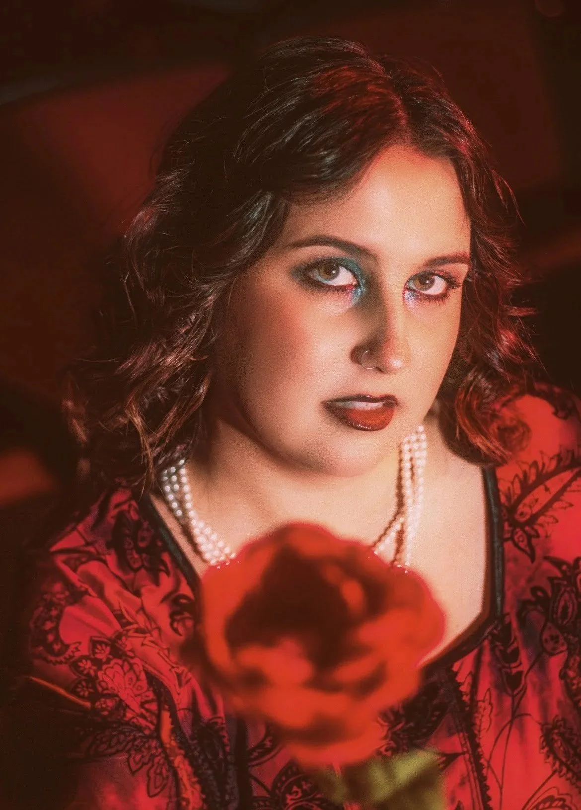 A woman with dark curly hair, piercings, eye makeup, and dark lipstick poses with a red rose in the foreground. She wears a red and black patterned garment and a pearl necklace, with a dark background.