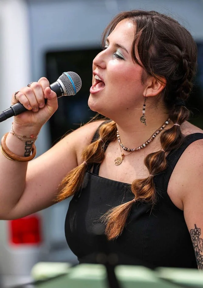 Woman singing into a handheld microphone, wearing a black sleeveless top and jewelry, with braided hair and tattoos on her arm.