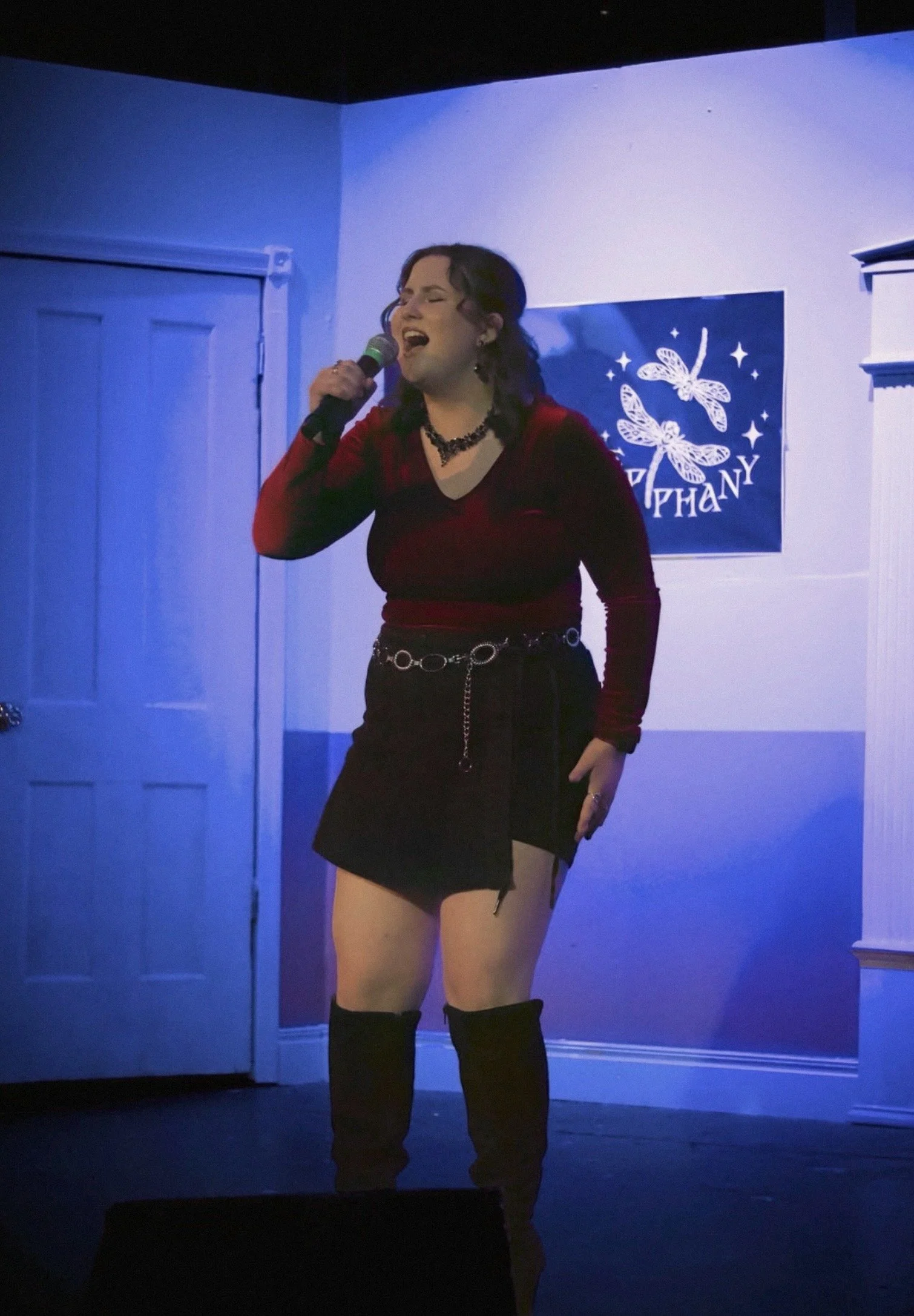A woman singing into a microphone on a stage with a blue background and starry poster.