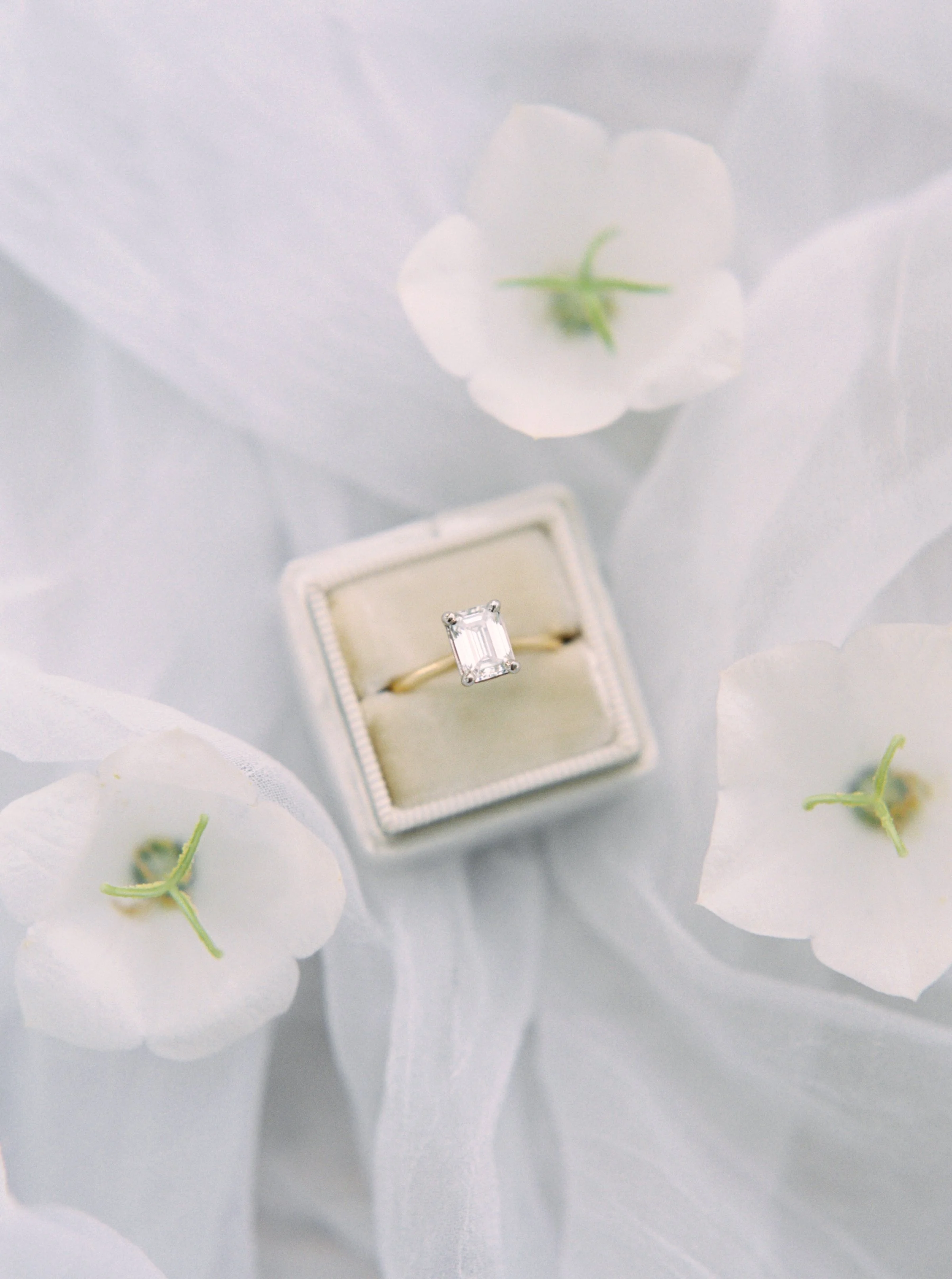 An engagement ring with a rectangular clear gemstone in a white jewelry box, surrounded by white flowers with pale green stems and leaves.