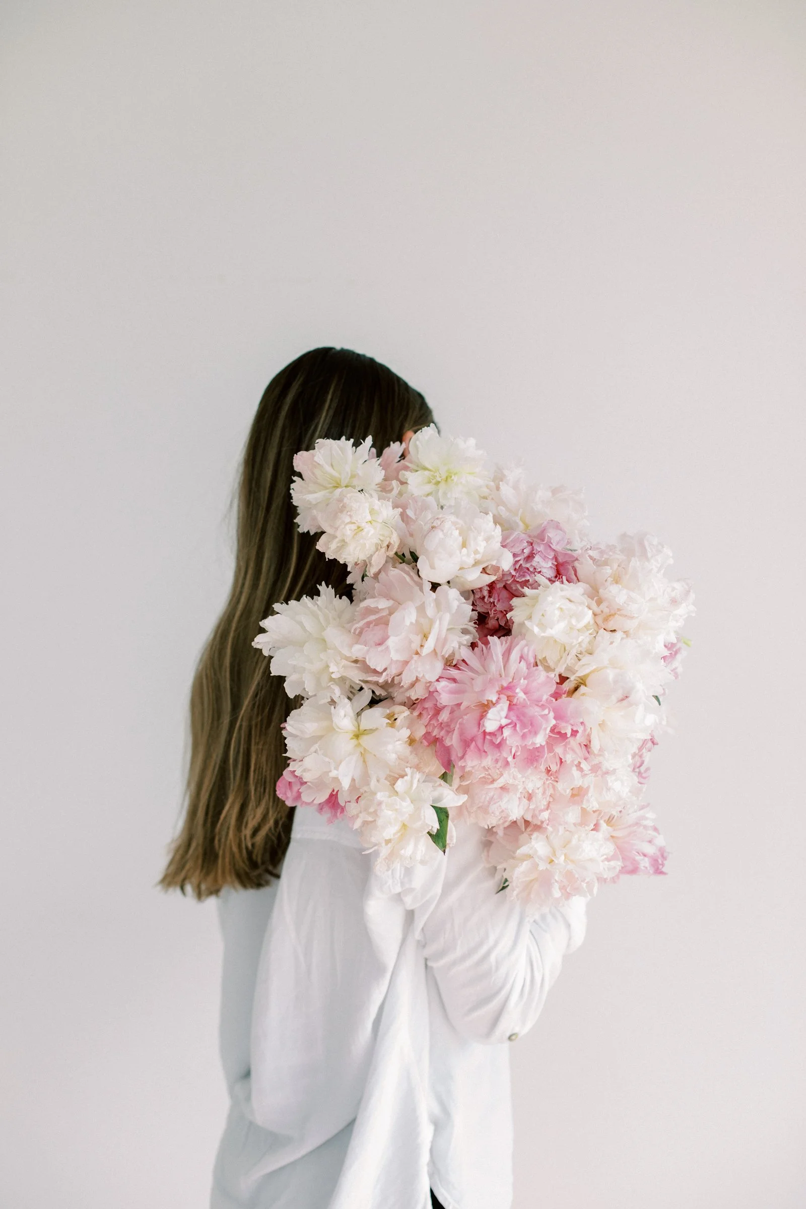 A woman with long brown hair wearing a white shirt is holding a large bouquet of soft pink and white peonies in front of her face, standing against a plain white background.