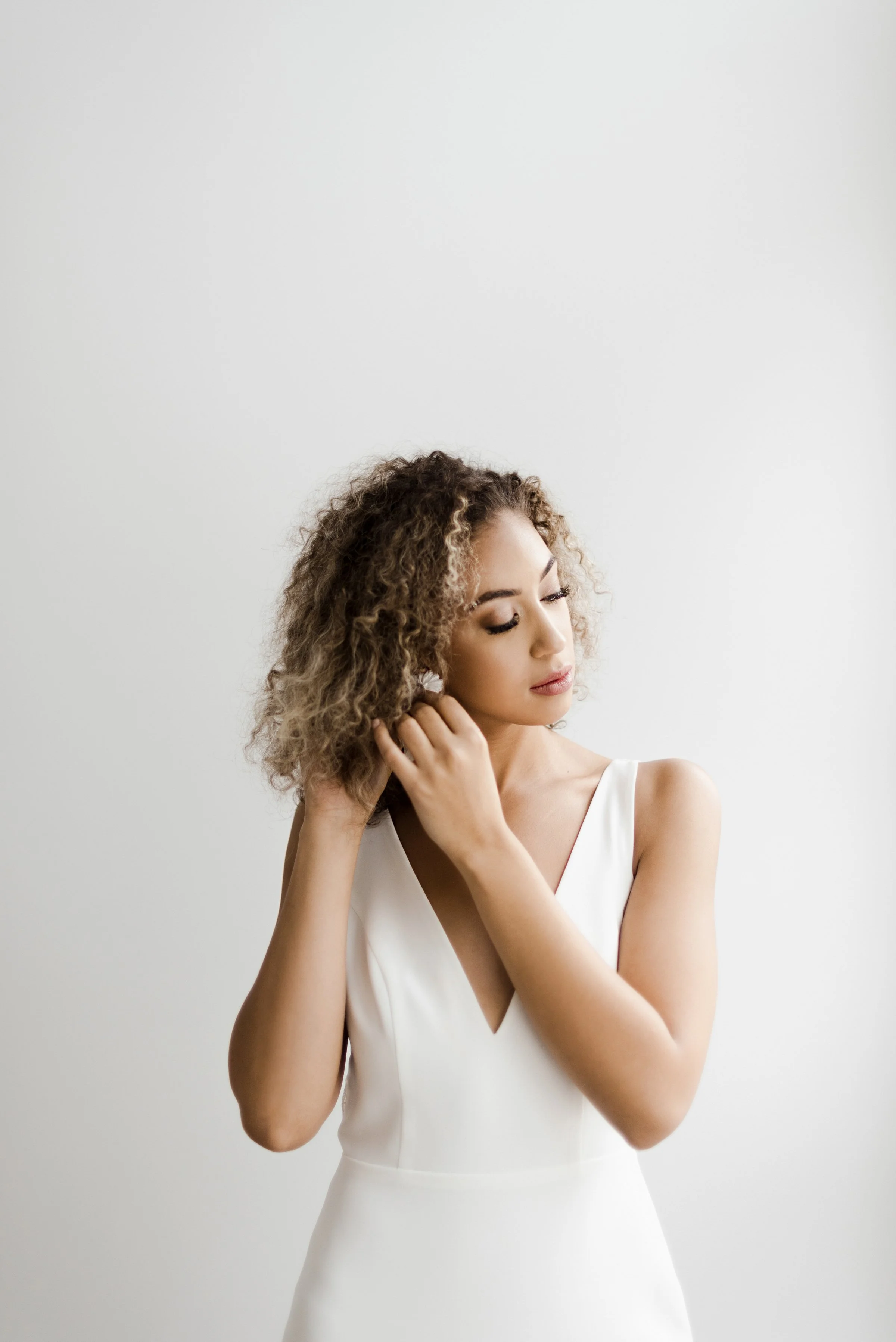 A woman with curly hair wearing a white sleeveless dress, standing against a plain white wall, gently touching her hair with one hand and looking down.