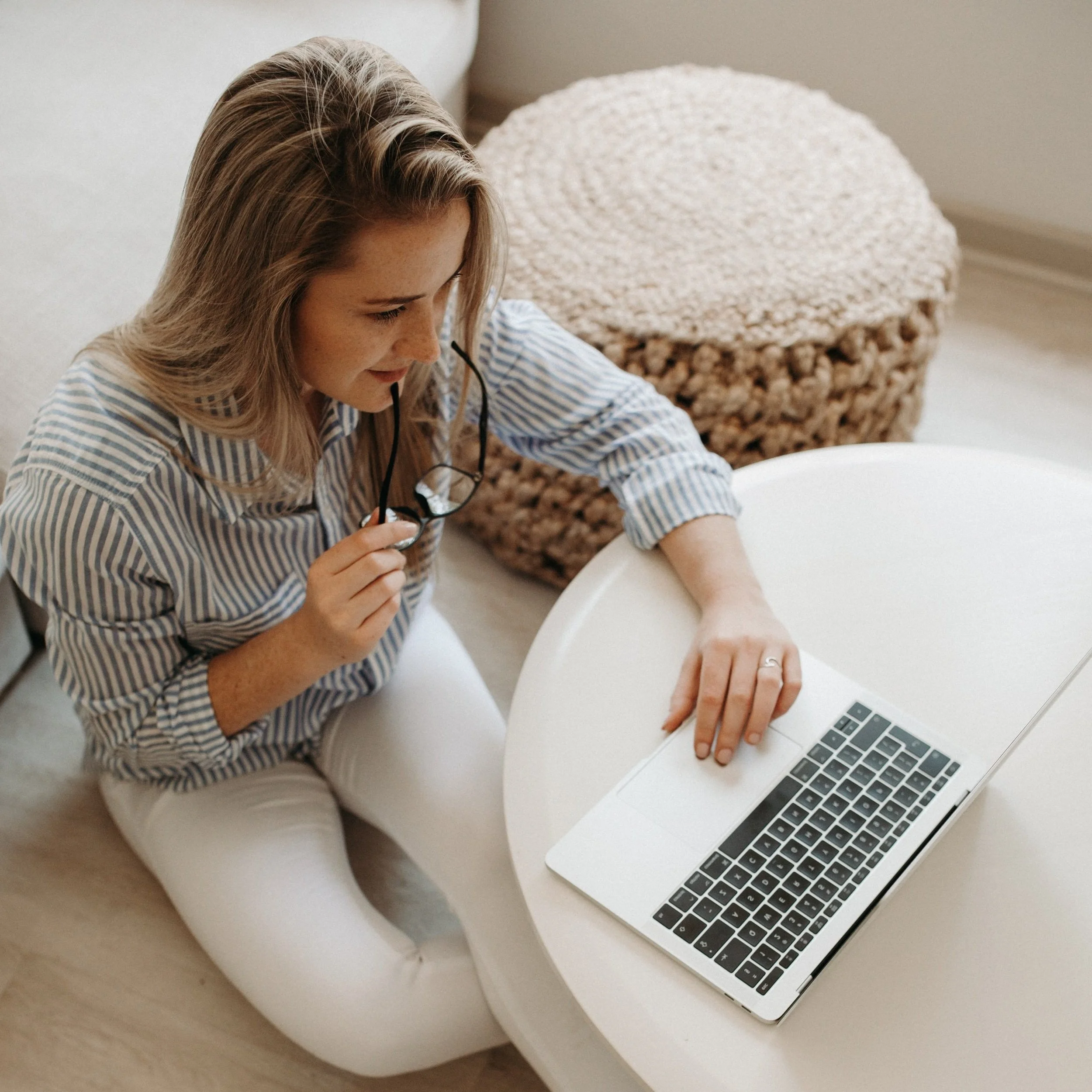 A woman with blonde hair in a striped shirt sitting at a white table, holding eyeglasses, with a laptop nearby and a woven basket in the background.