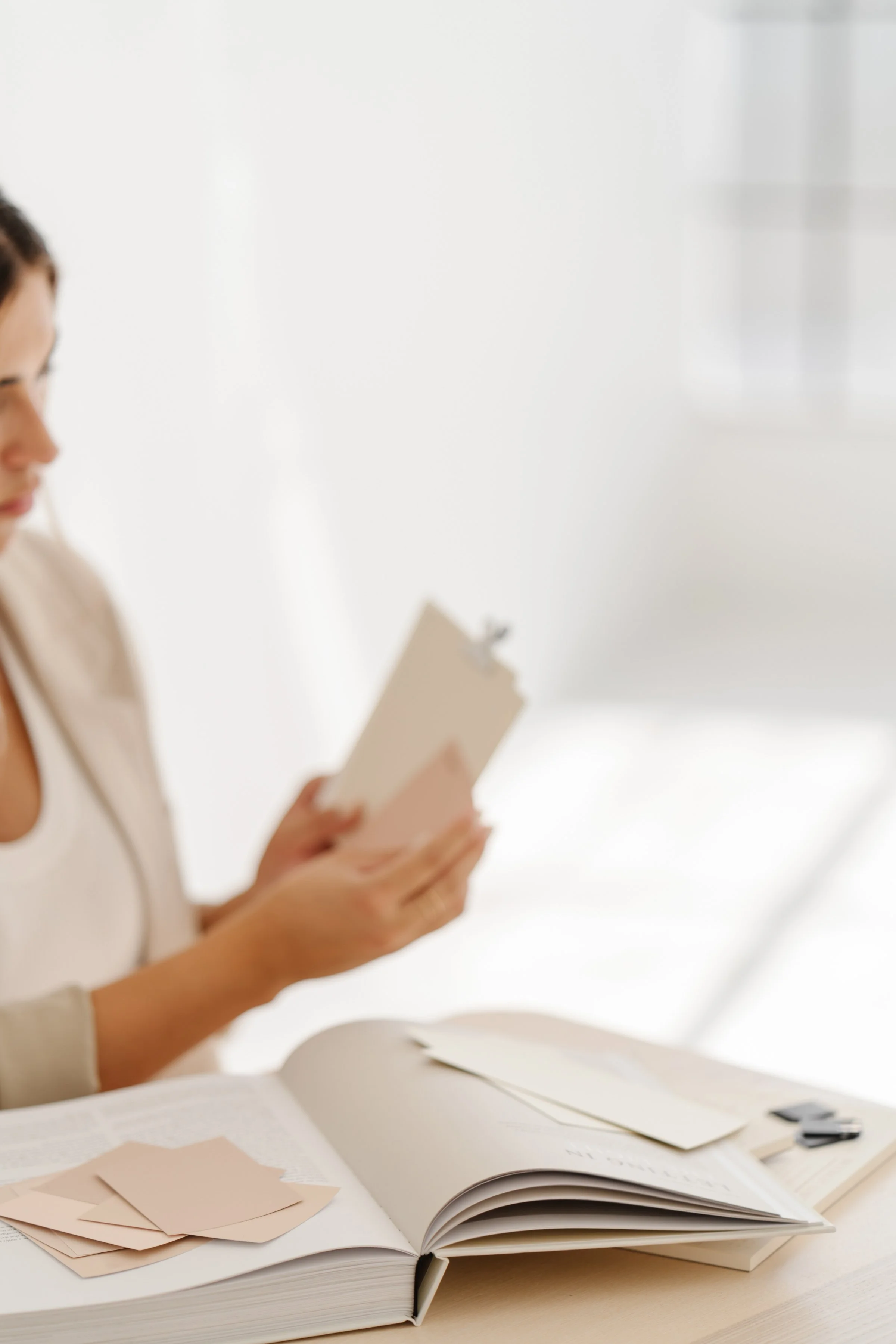 A person sitting at a white desk with an open large book, holding a card, with color swatches and paper scraps spread around.