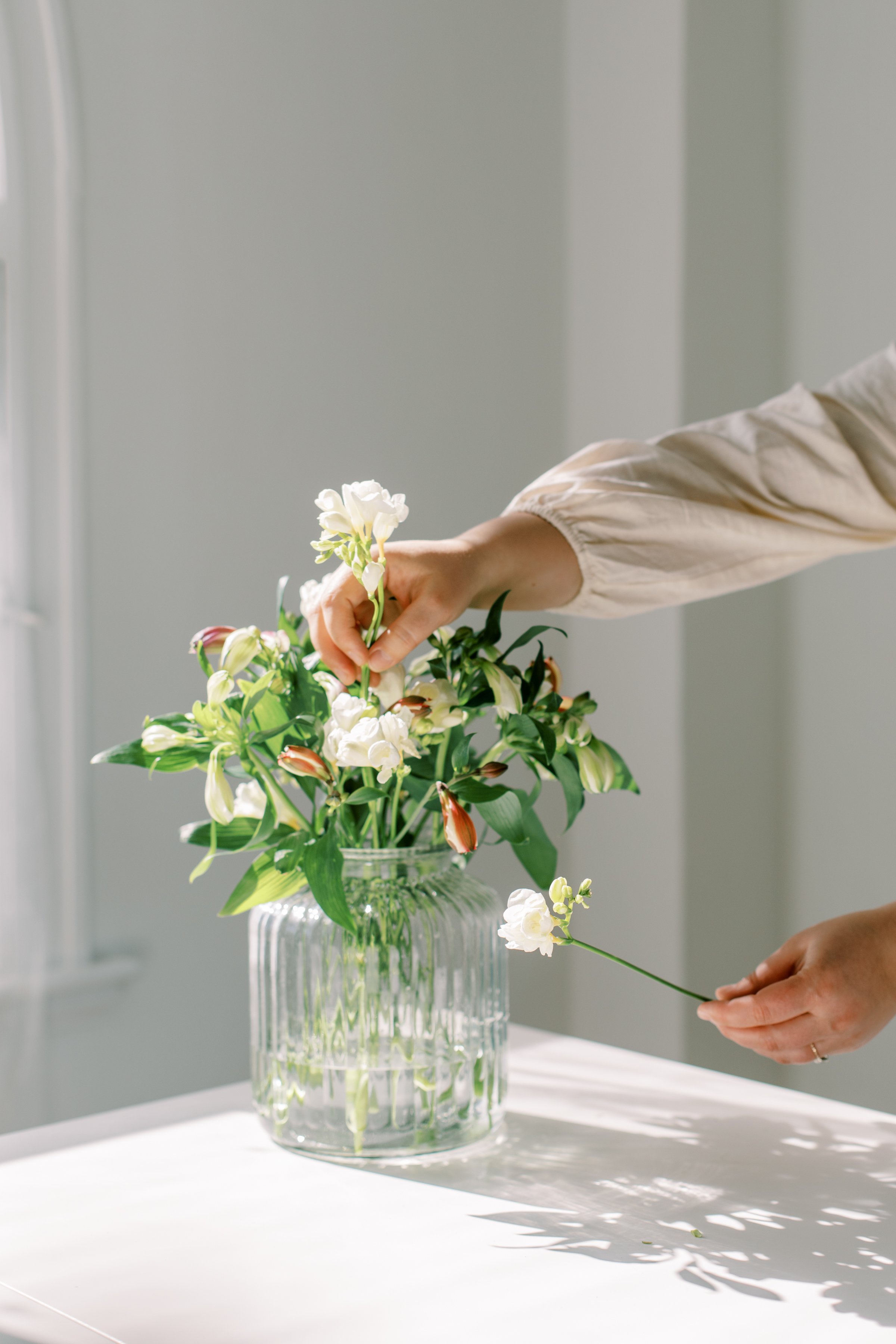 Person arranging a fresh flower bouquet in a clear glass vase on a white table, with natural sunlight casting shadows.