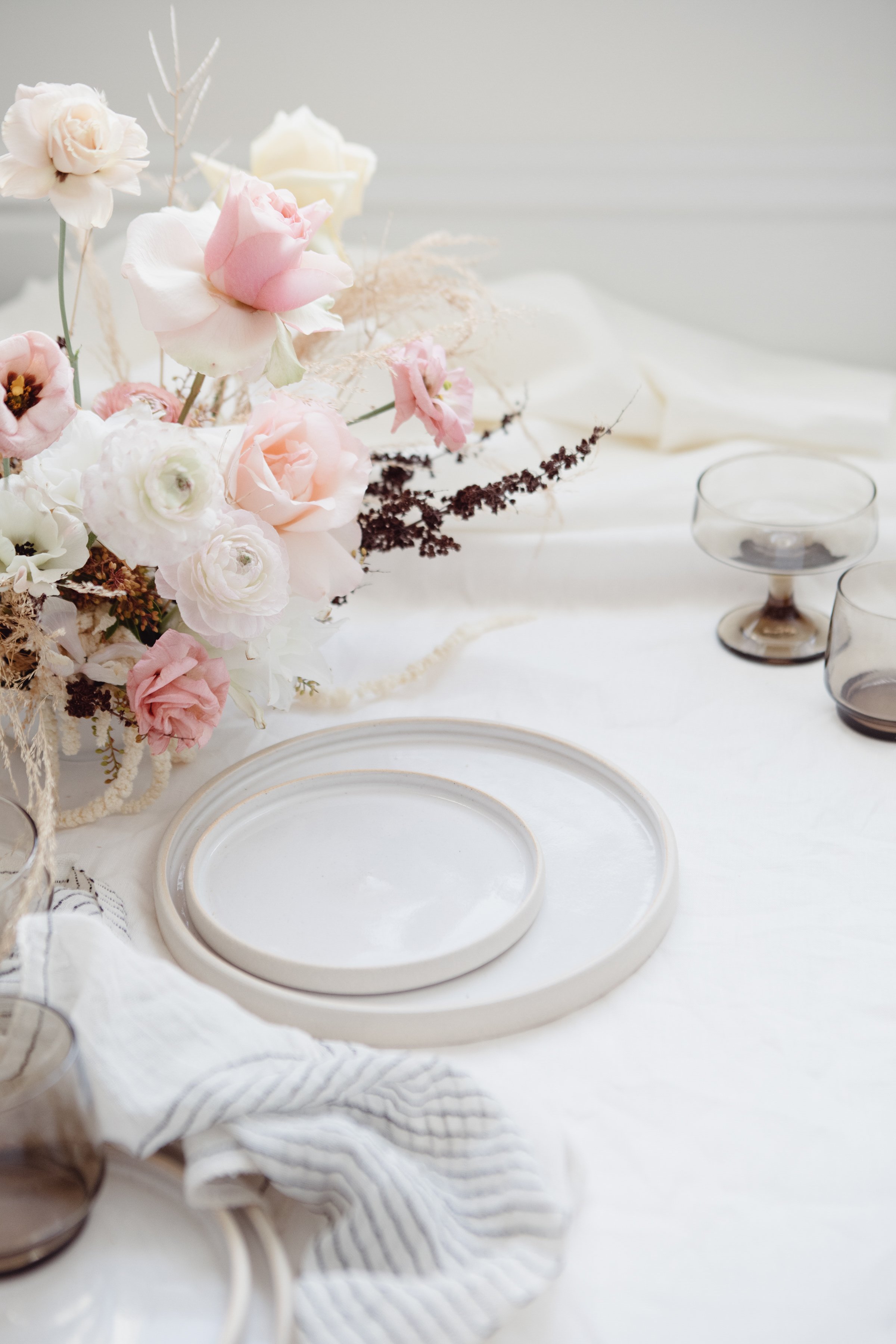 A floral centerpiece with pink and white roses and dried flowers on a dining table set with empty white plates and glassware.