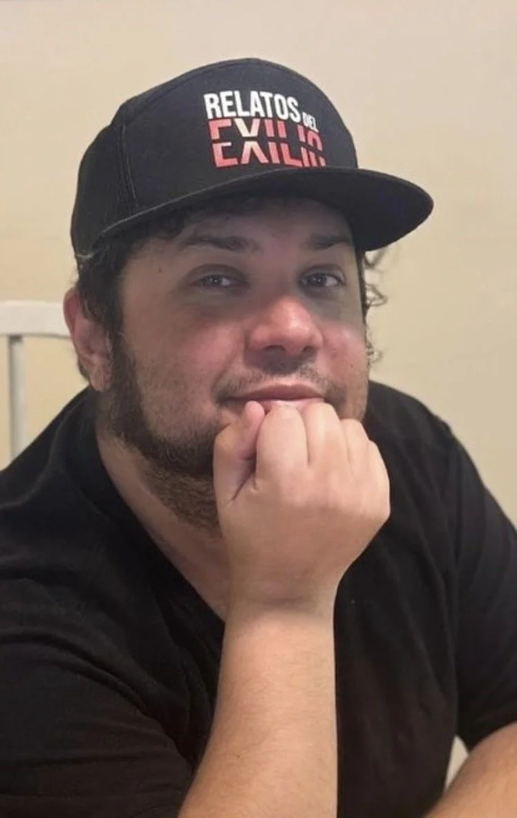 A man wearing a black baseball cap with the words 'RELATOS DEL EXILO' in white and pink, and a black shirt. He has dark hair, a beard, and is resting his chin on his hand while looking at the camera.