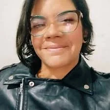 A woman with glasses, dark hair, and a black leather jacket, smiling.