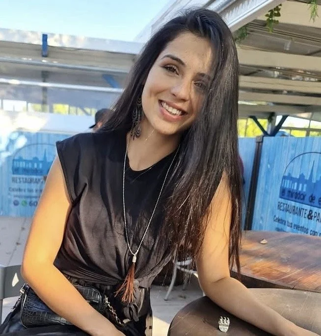 Smiling young woman with long dark hair, wearing a black sleeveless top, sitting outdoors at a wooden table.