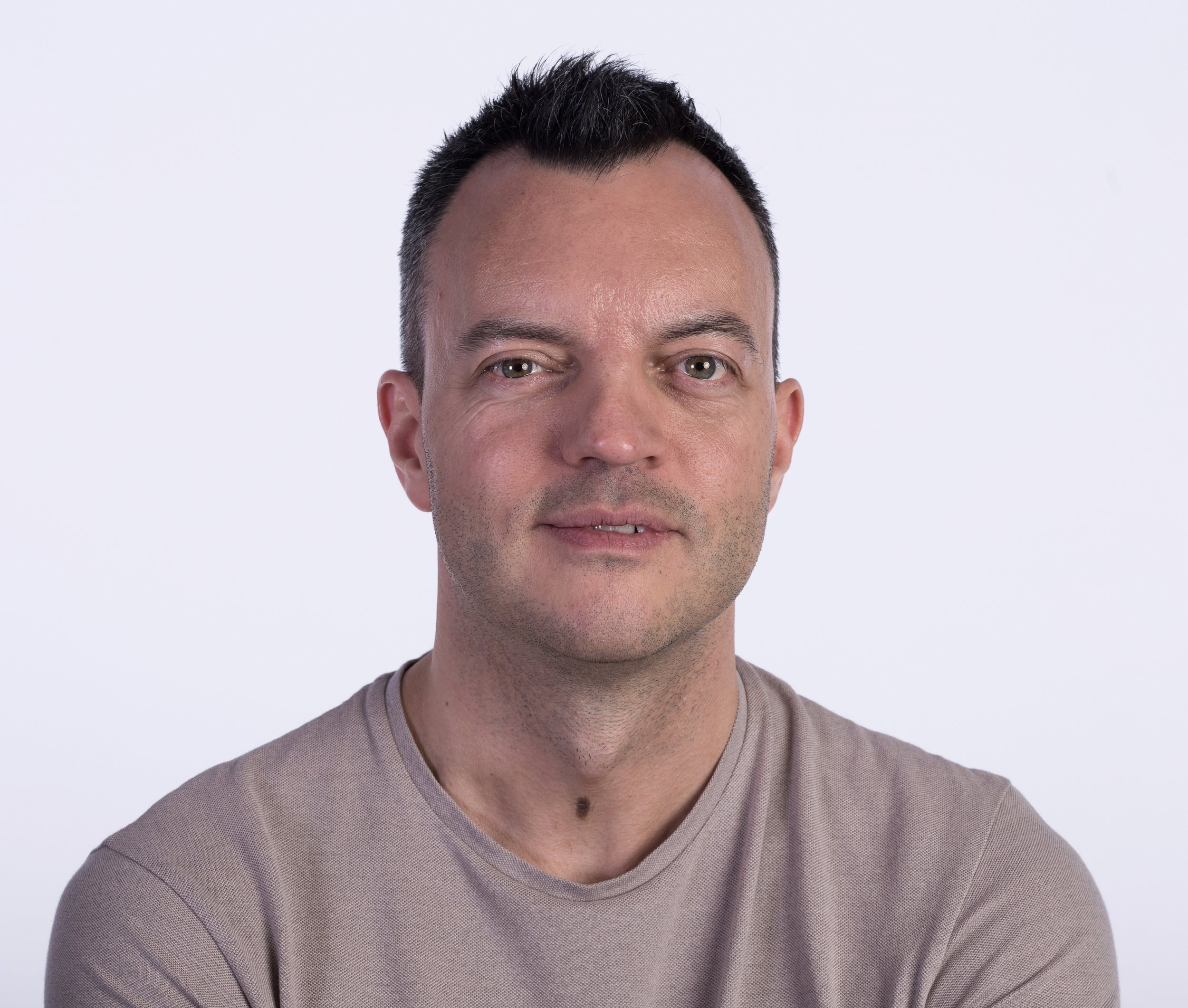 Close-up of a man's face with short dark hair, green eyes, and a slight smile, wearing a beige V-neck shirt against a plain white background.