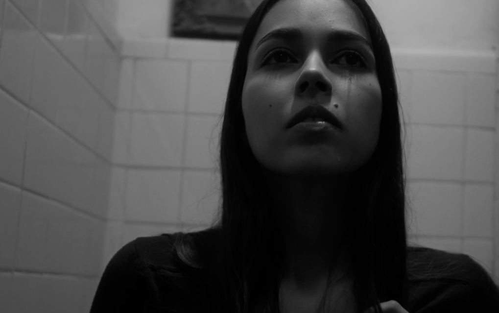 Black and white photo of a young woman with long dark hair, ignoring the camera, in a tiled room.