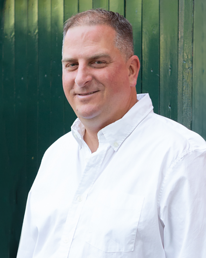 A smiling man in a white button-up shirt standing in front of a green wooden fence.