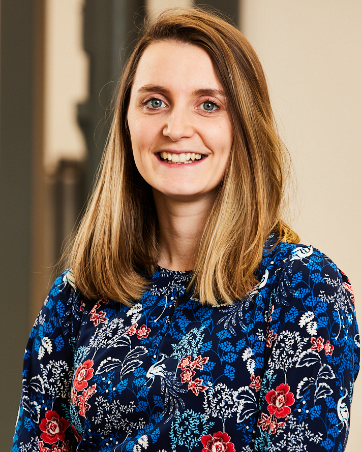 A young woman with long, straight, light brown hair and blue eyes smiling at the camera, wearing a colorful floral blouse.