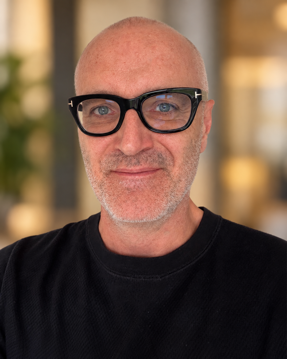 A smiling man with a bald head, wearing large black glasses with a white logo, and a black shirt. The background is blurred indoors.