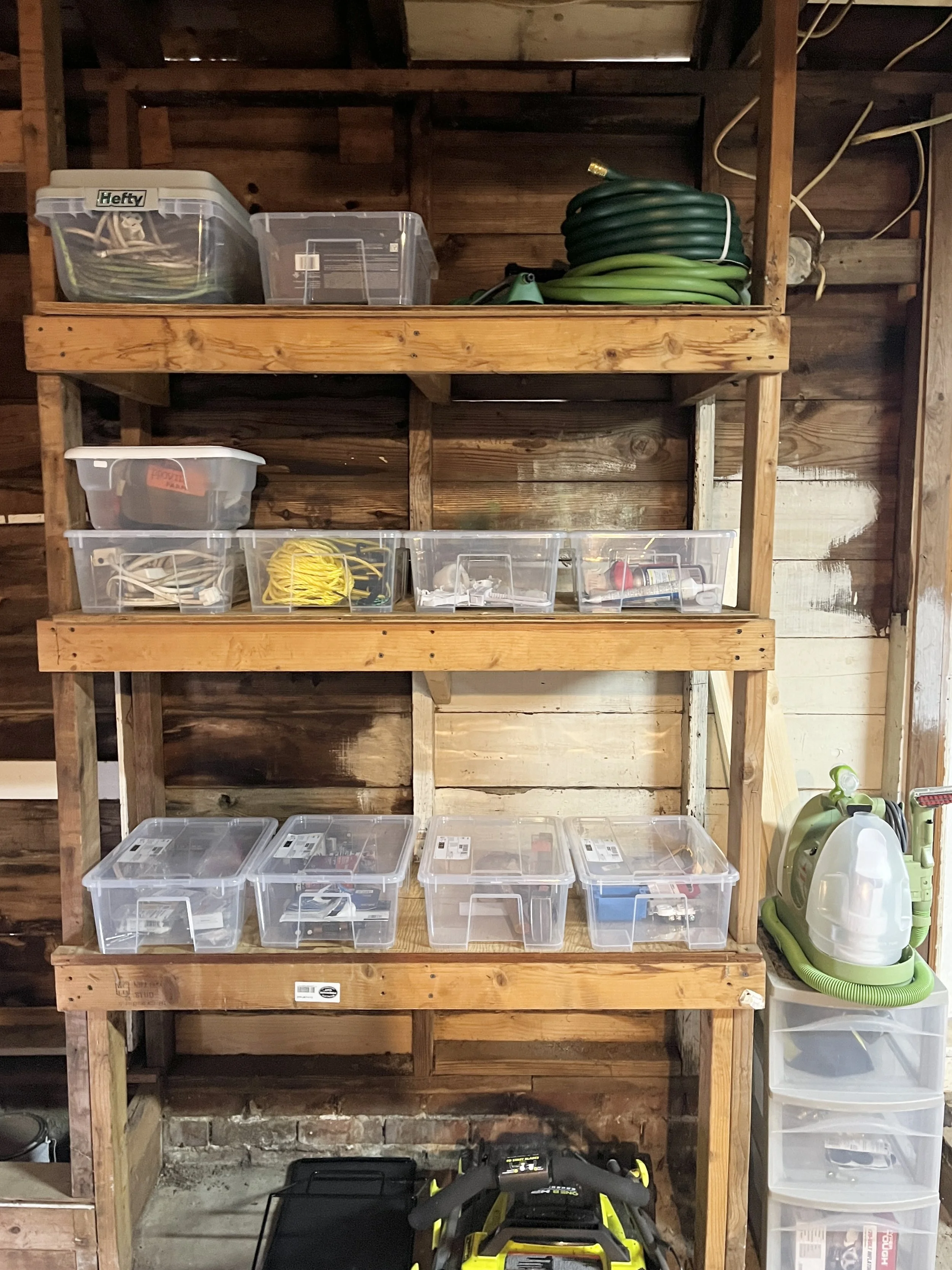 Wooden garage shelving unit with clear plastic storage bins, a green garden hose, and a vacuum cleaner.