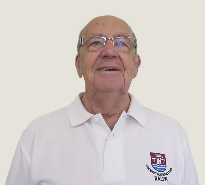 An elderly man wearing glasses and a white collared shirt with a logo that says "Short Mat Bowls Club RALPH".