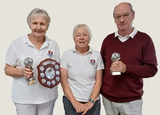 Three elderly people, two women and one man, standing against a plain background. The woman on the left is holding a trophy and a shield-shaped award. The woman in the middle is standing with her hands together, and the man on the right is holding a trophy. All three are smiling and wearing casual clothing with two of them in white polo shirts featuring a crest.