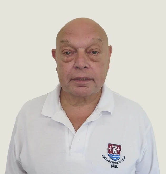 An older man with a bald head and droopy eyes wearing a white polo shirt with a crest on the left side.