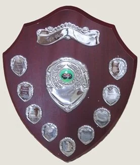 A wooden shield-shaped trophy plaque with a silver center and ten smaller silver plates around it, some with decorative engravings, and a silver holder at the top.