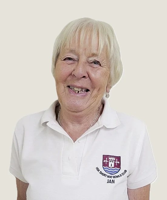 Senior woman smiling, wearing a white polo shirt with a logo and the name Jan, against a plain background.