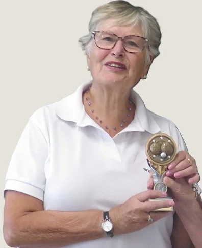 An elderly woman with short gray hair and glasses, wearing a white polo shirt, holding a small model of a human brain and spinal cord.