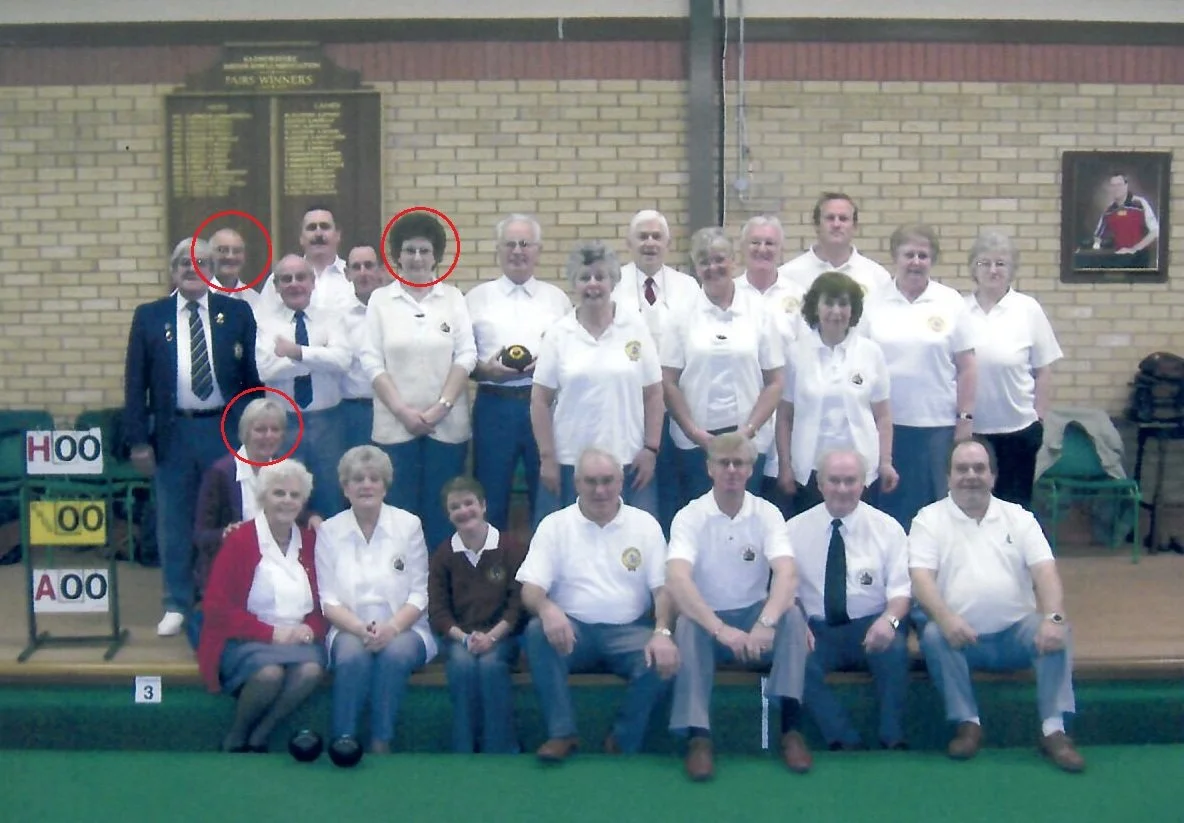 Group of 16 people posing for a photo in an indoor setting, some wearing white shirts with a badge, others in casual clothing, with a brick wall background and framed portraits on the wall.