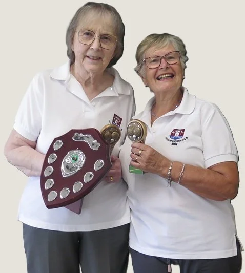 Two elderly women in white polo shirts holding a shield-shaped trophy and a pair of round awards, smiling.