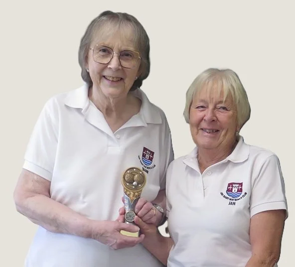 Two elderly women wearing white polo shirts with embroidered logos, standing close together and smiling at the camera. One woman is holding a small gold-colored trophy, and both women seem happy.