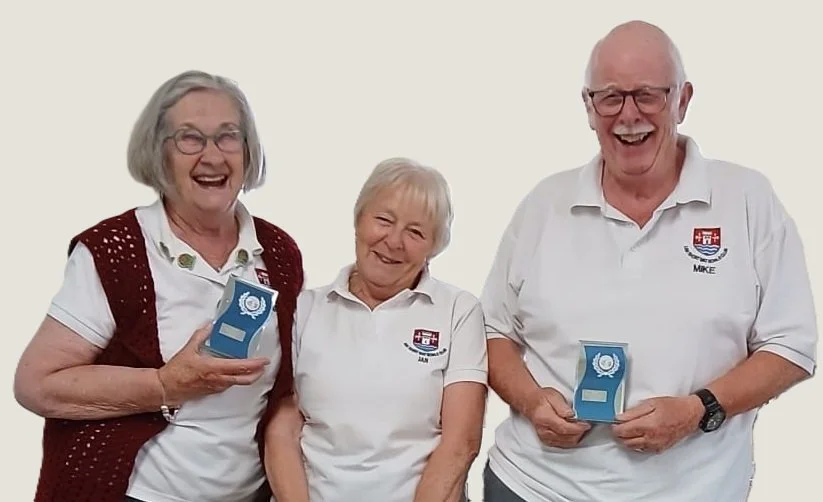 Three smiling elderly people standing together, two women and one man, wearing white polo shirts with a crest, holding awards, against a plain background.