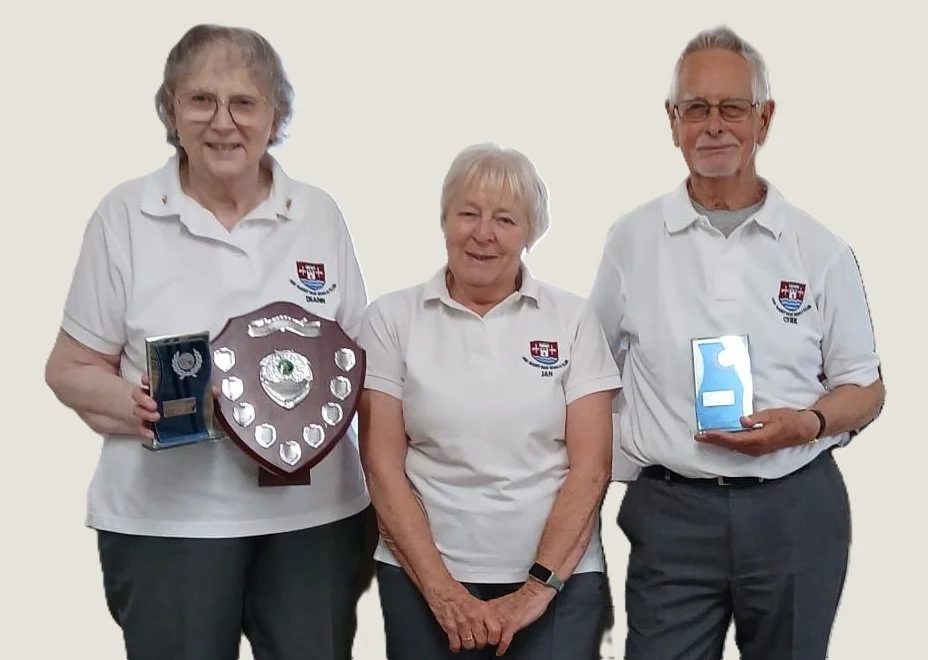 Three elderly individuals wearing matching white shirts with a logo, standing together, holding awards, and smiling.