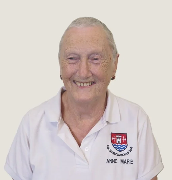 An elderly woman with short gray hair wearing a white polo shirt with a crest and the name Anne Marie, smiling against a plain background.