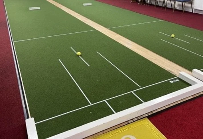 Indoor mini-golf putting game with a green turf, white lines, two yellow golf balls, and a partial view of chairs and a red carpet.