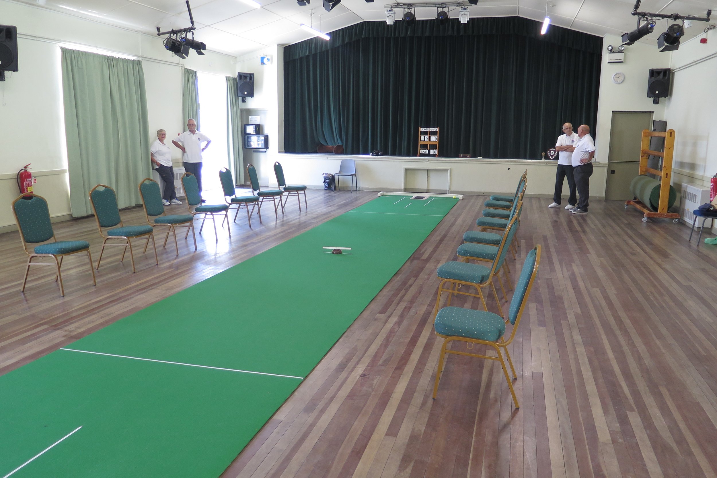 An indoor activity room with a green indoor golf putting area, chairs arranged along the sides, and four people standing near the back wall. The room has a stage with a dark green curtain, multiple lights and speakers, and a scoreboard on the stage. There are windows with green curtains and a fire extinguisher in the corner.