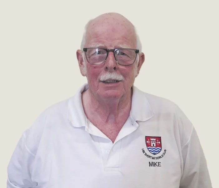 An elderly man with glasses, white hair, and a mustache, wearing a white polo shirt with a logo that reads 'U.S. Short Mat Bowls Club' and the name 'Mike' on it, standing against a plain background.