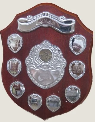 A wooden shield-shaped plaque with multiple silver medals attached, featuring year and event engravings, and a larger central medal with intricate designs.