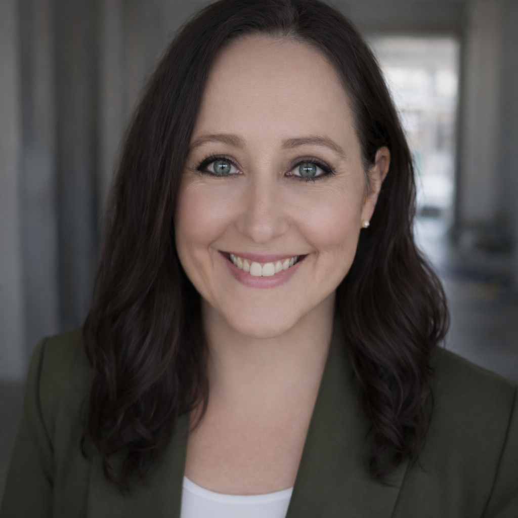 A woman with shoulder-length dark brown hair, light skin, blue eyes, and a bright smile, wearing a dark green blazer and a white top, standing indoors with a blurred background.