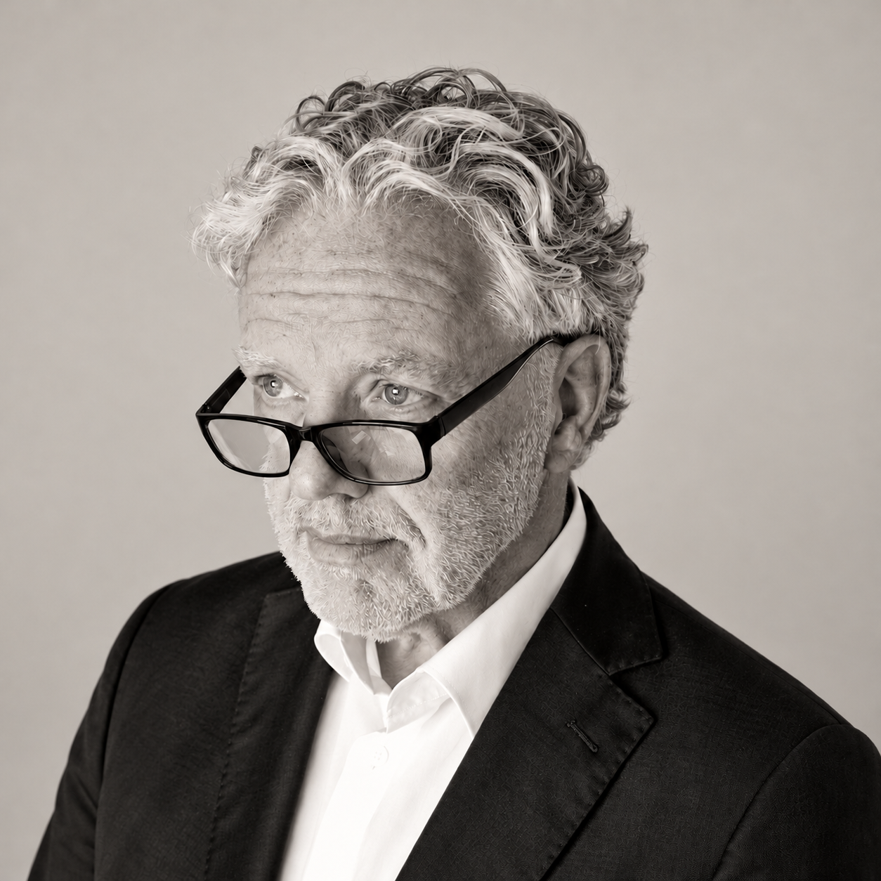 Black-and-white portrait of an older man with curly gray hair and glasses, wearing a suit and white shirt, looking to the side.